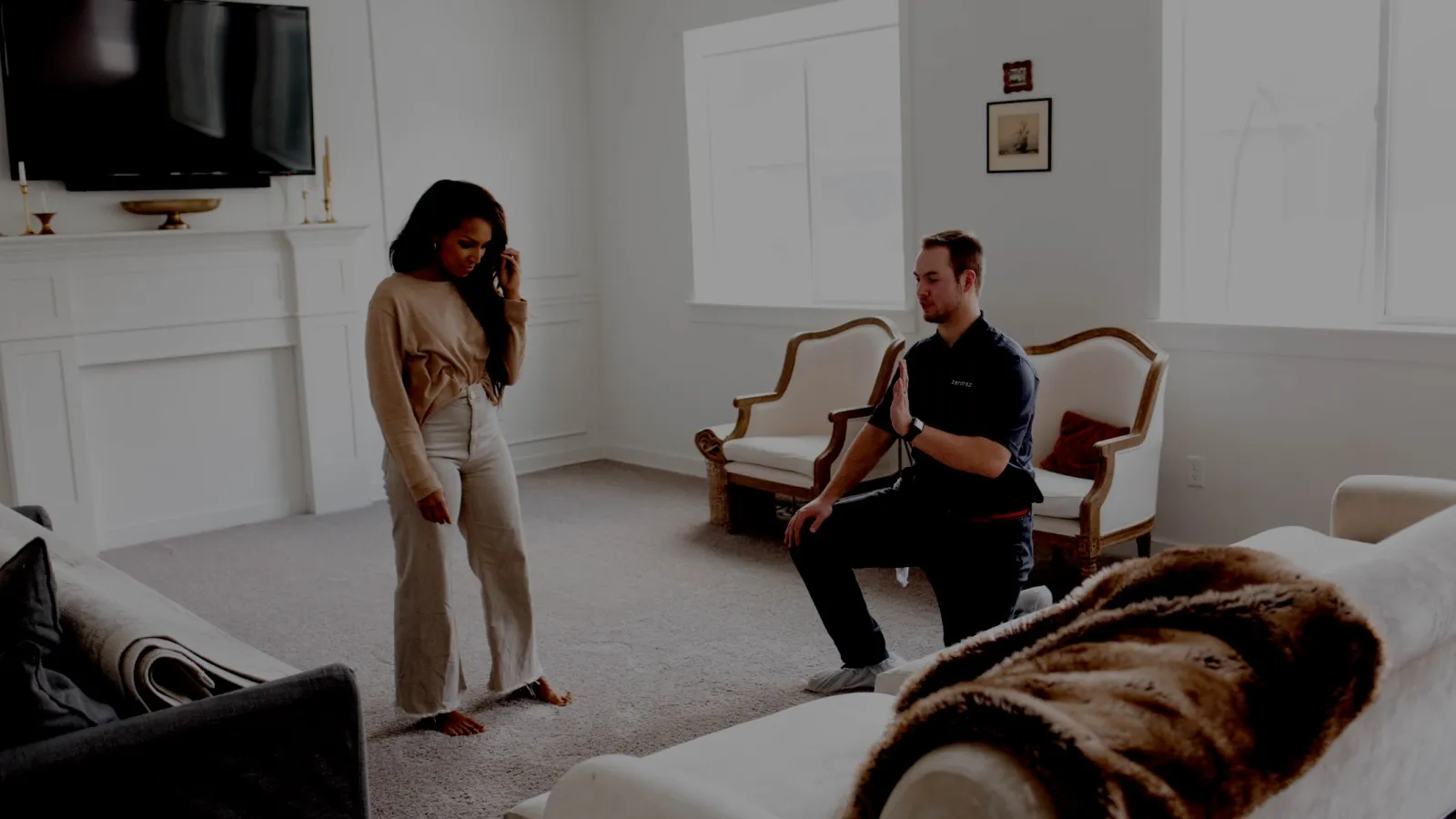 Man kneeling and gesturing while woman stands in a bright living room with couches and a TV