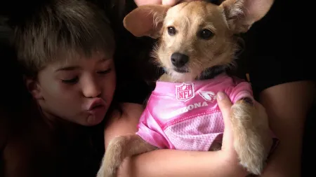 A girl and boy hold a small dog in NFL jersey against a dark background, showcasing their playful bond.