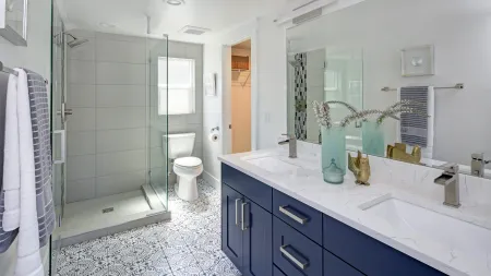 Modern bathroom with patterned tile floor, glass shower, white toilet, blue vanity, and double sink with mirror.