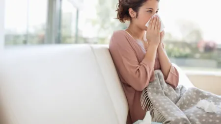 A woman sitting on a couch, looking distressed with tissues in hand, wrapped in a cozy blanket.