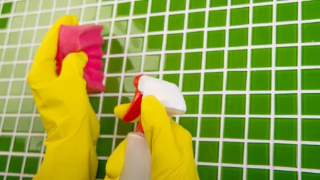 Hands wearing yellow gloves cleaning green tiled wall with a pink sponge and spray bottle.