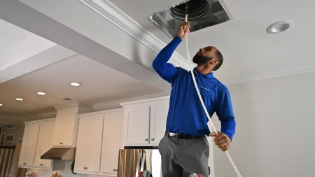 Technician in blue shirt inspecting and cleaning a kitchen air vent on ceiling with a brush and ladder.