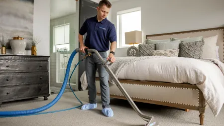 Professional cleaner using steam cleaning machine on bedroom carpet in modern home with beige decor.