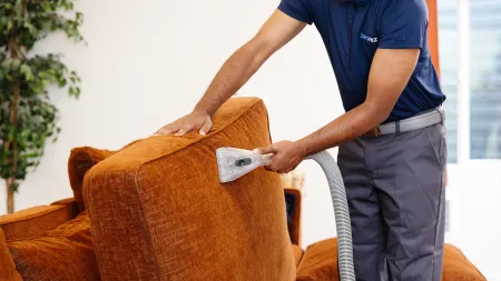 Man in uniform professionally cleaning orange sofa upholstery with vacuum extractor in bright living room