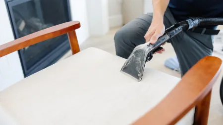 Professional cleaning service vacuuming beige fabric chair upholstery with specialized tool near fireplace.