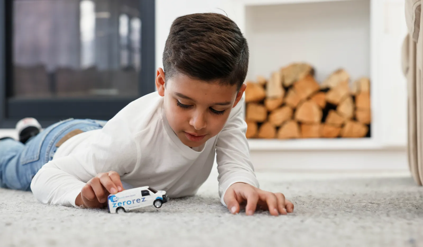 Young boy lying on carpet playing with a toy van labeled Zerorez near a fireplace with stacked firewood.
