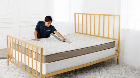 Person adjusting a mattress on a modern bed frame in a bright, minimalistic bedroom setting.